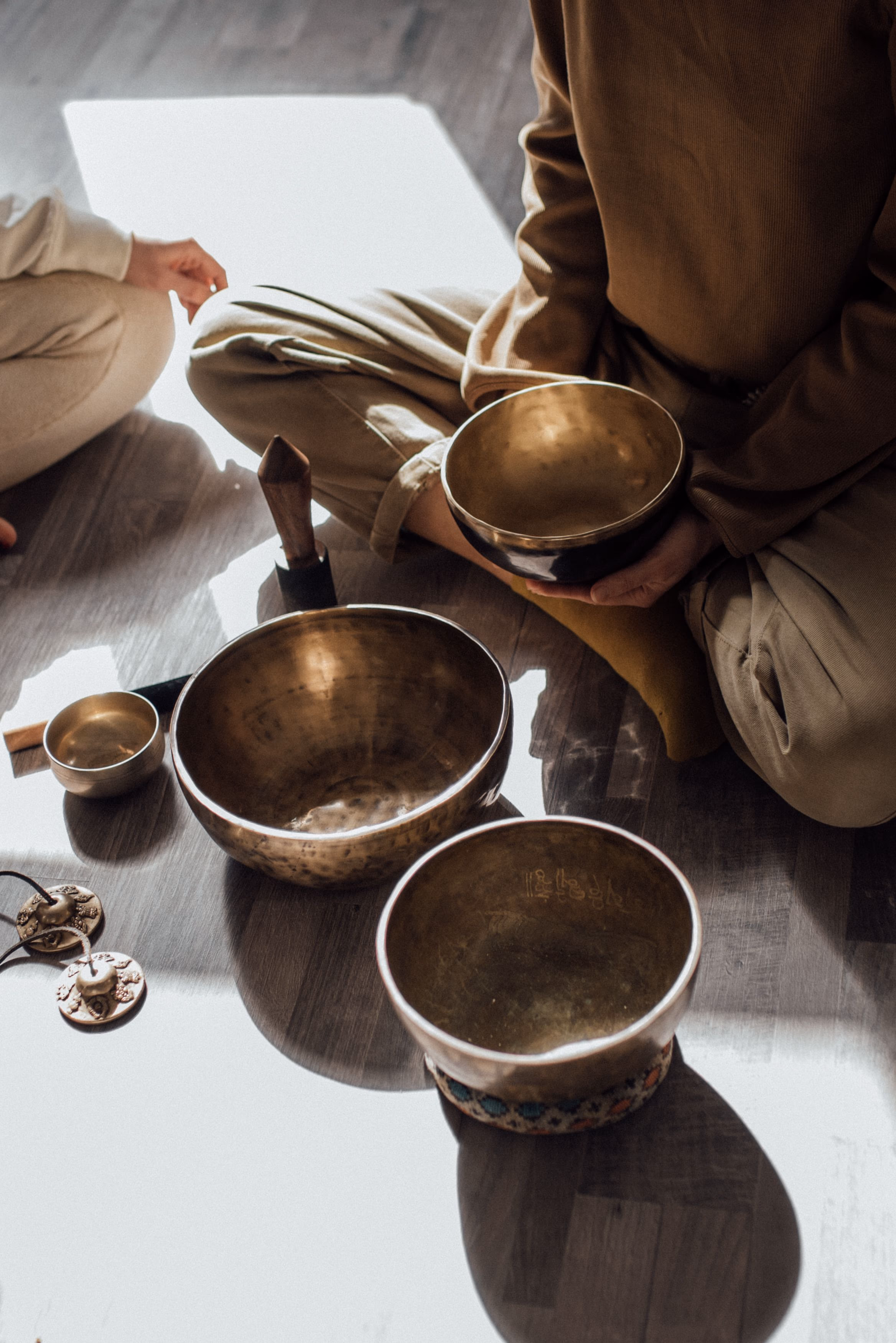 Two people sitting on a warm floor with brass singing bowls between them.