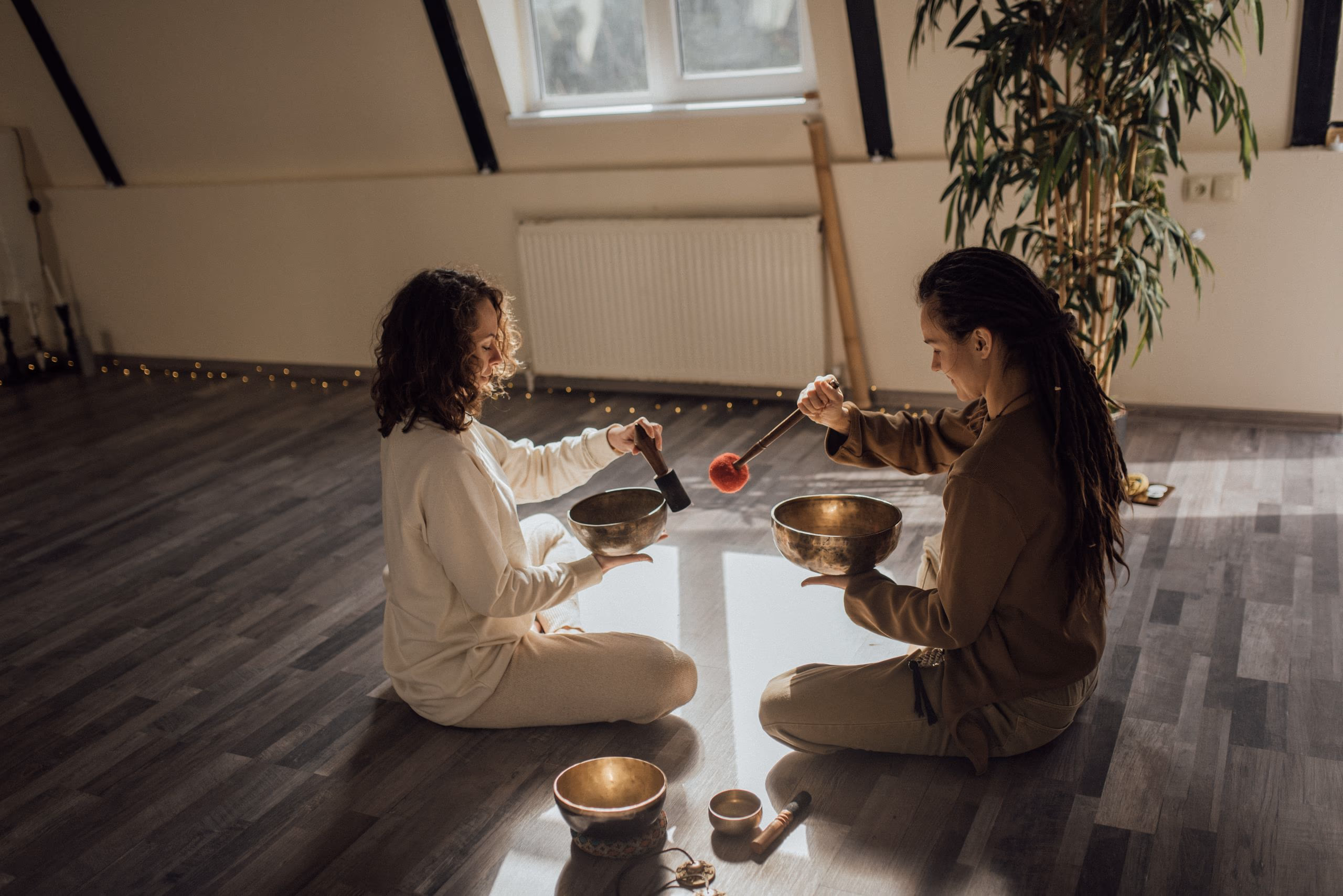 Two people sitting facing each other on a warm wooden floor, each holding a brass singing bowl.