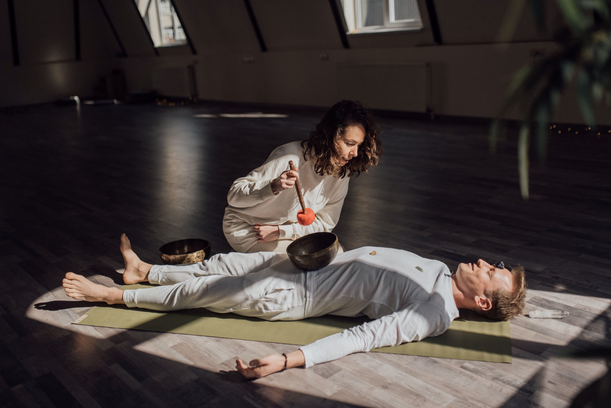 A practitioner kneeling beside someone lying on a mat, holding a singing bowl above them.