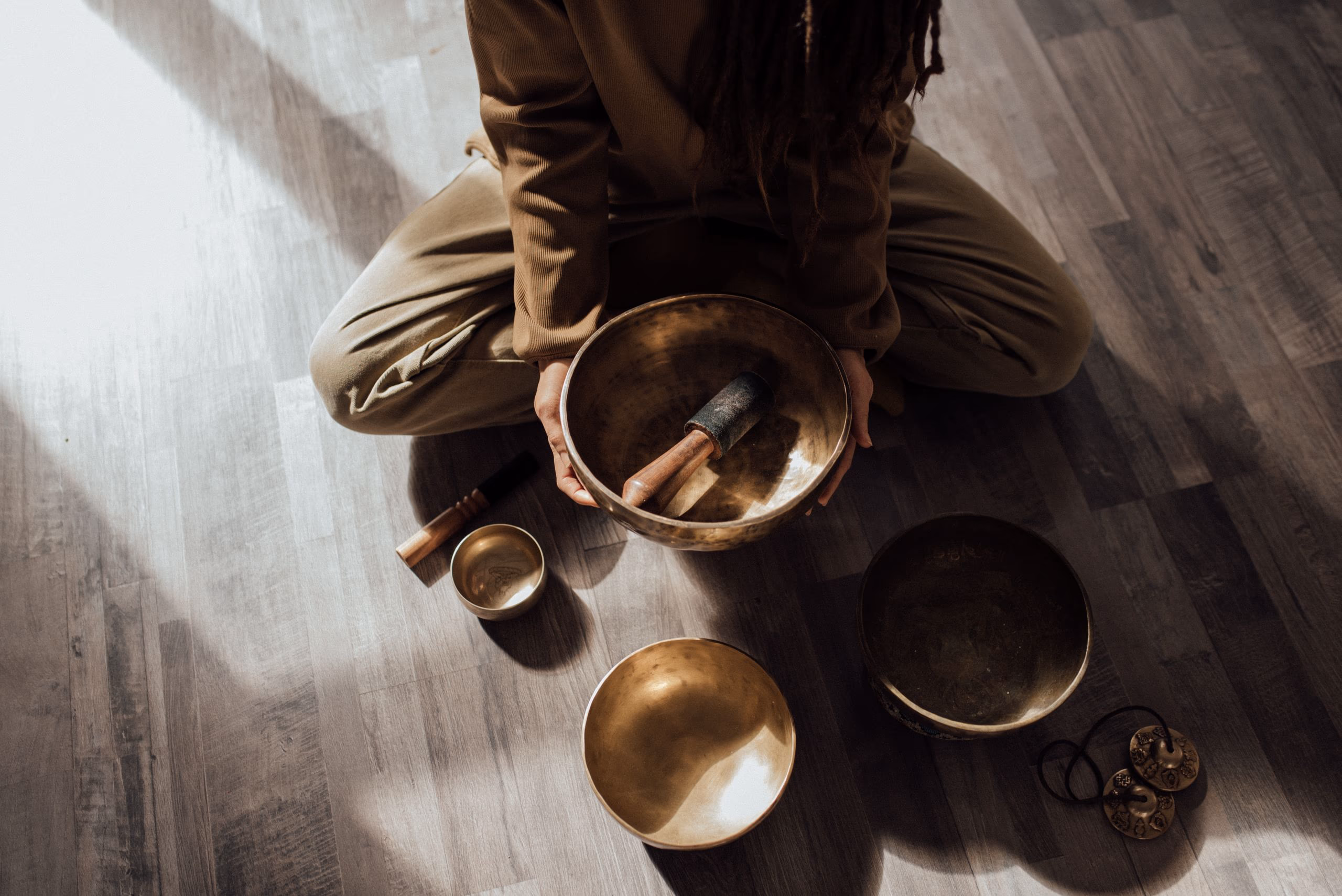 Seen from above, a person sitting on a warm oak floor surrounded by brass singing bowls in a shaft of sunlight.